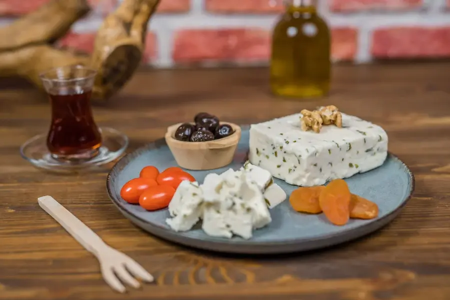 “Otlu peynir, zeytin, kuru kayısı ve domatesle birlikte servis edilmiş geleneksel kahvaltı tabağı – yanında çay bardağı ve doğal zeytinyağı şişesiyle sunum.”